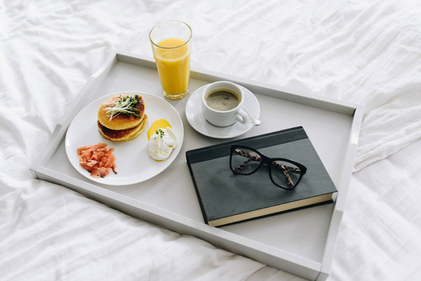 Café da manhã nutritivo e saboroso na cama na bandeja com café, suco de laranja, óculos e livro
 - Foto, Imagem