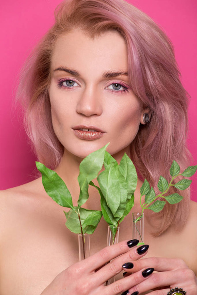 Shiny golden make-up on a beautiful young European model girl holding a twig with green leaves in hand isolated on an pink background in the photo studio - Fotografie, Obrázek