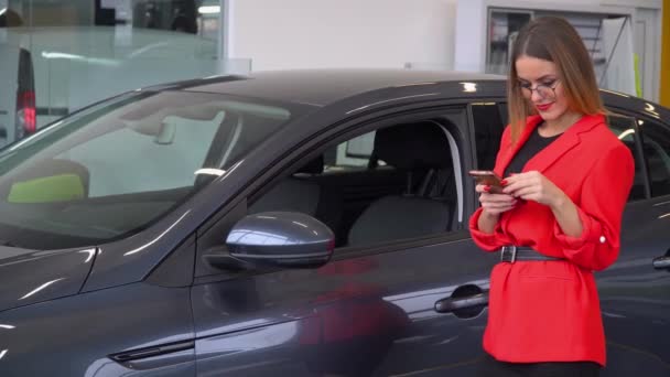 Beautiful woman uses smartphone while sitting in a car at a car dealership - Materiaali, video