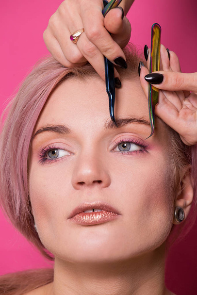 Close-up of a beautiful girl of European appearance holds in her hand tweezers for eyebrows near the face in the studio isolated on a pink background - Zdjęcie, obraz