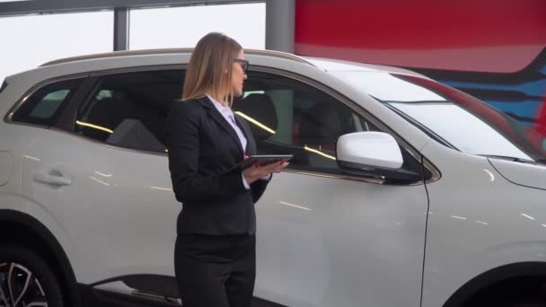 The female seller stands by the car in the car dealership and makes notes on the tablet - Video