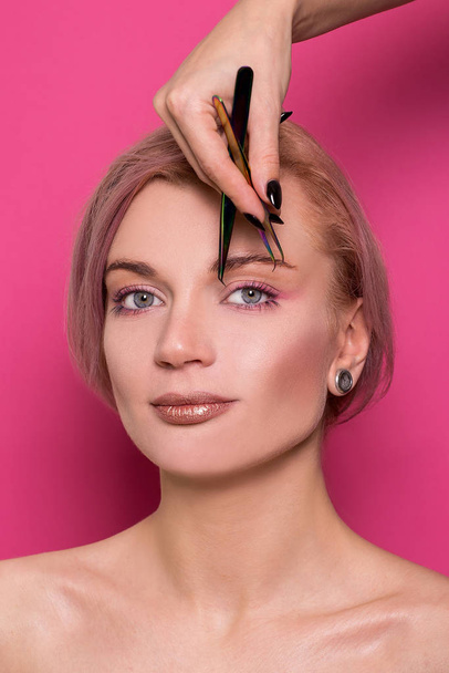 Close-up of a beautiful girl of European appearance holds in her hand tweezers for eyebrows near the face in the studio isolated on a pink background - Fotografie, Obrázek
