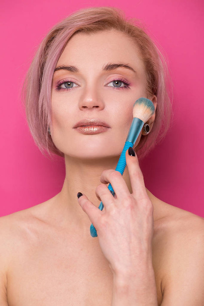 Beautiful blonde girl of European appearance holds a makeup brush in her hand near her face isolated on a pink background - Фото, зображення