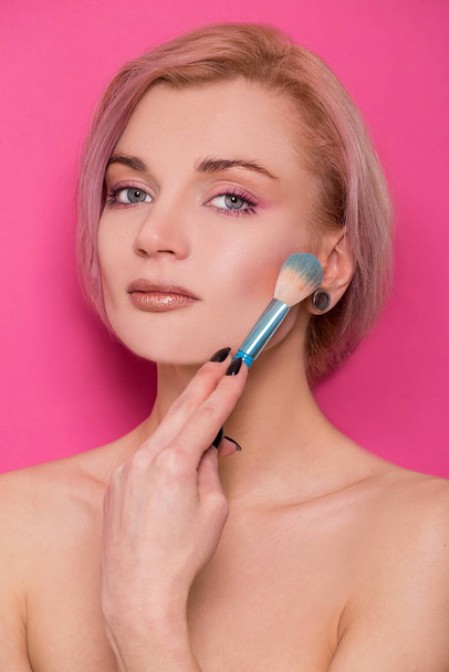 Beautiful blonde girl of European appearance holds a makeup brush in her hand near her face isolated on a pink background - Valokuva, kuva