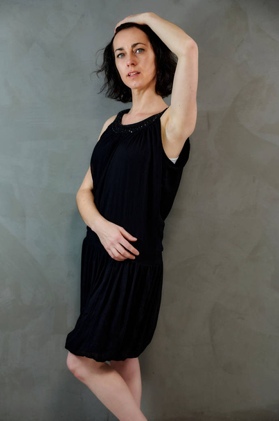 Full length portrait of a pretty emotional brunette woman in black dress on alternative gray background in vintage interior. Standing right in front of the camera. - Fotografie, Obrázek