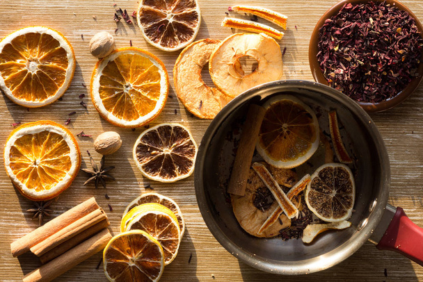 Top view of dry pieces of lemon, orange and apple, cinnamon sticks, bay leaves, nutmegs, star anise and dry cutted hibiscus as ingredients for tea and the saucepan - Photo, Image