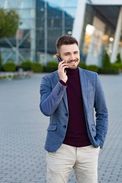 Portrait of handsome smiling man in casual wear holding smartphone. Successful manager makes a call. Young businessman talking on the phone, standing on city street near business center. - Foto, Bild