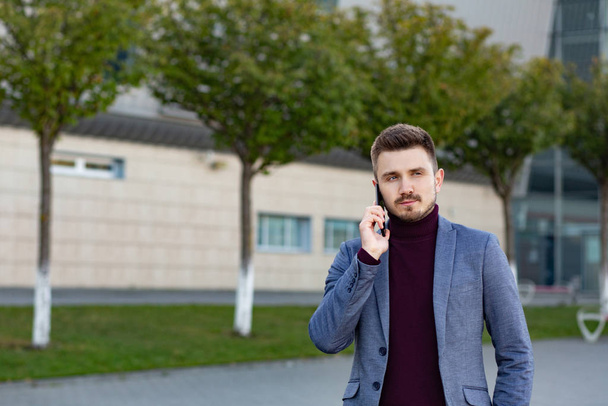 Portrait of handsome serious man in casual wear holding smartphone. Successful manager makes a call. Young businessman talking on the phone, standing on city street near business center. - Photo, image
