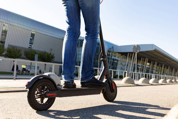 Πόδια ενός άνδρα που στέκεται σε e-scooter σταθμευμένα στο πεζοδρόμιο στο cityscape στο ηλιοβασίλεμα. Trendy αστικές συγκοινωνίες με σύγχρονο ηλεκτρικό σκούτερ. Οικολογική έννοια της κινητικότητας. - Φωτογραφία, εικόνα