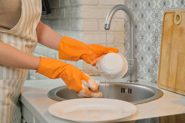 Jovem mulher em luvas de avental lavar pratos com esponja e detergente
 - Foto, Imagem