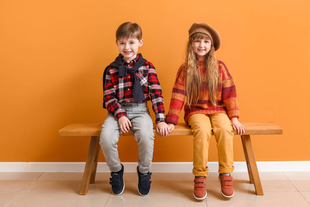 Lindos niños pequeños en ropa de otoño sentados en el banco cerca de la pared de color
 - Foto, Imagen