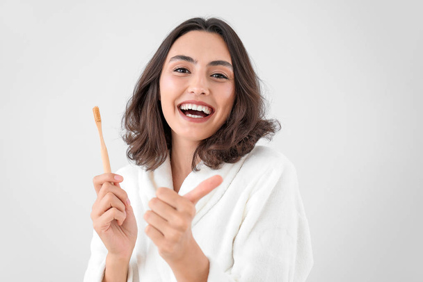 Beautiful young woman with toothbrush showing thumb-up gesture on light background - Φωτογραφία, εικόνα