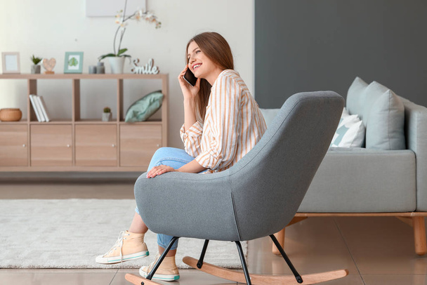 Piękna kobieta rozmawia przez telefon komórkowy siedząc w fotelu w domu - Zdjęcie, obraz