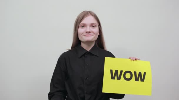 Happy charming blonde woman keeping advertising paper WOW with yellow wall - Záběry, video