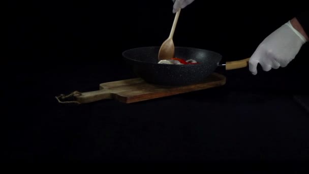 Σεφ αναδεύεται λαχανικά σε ένα τηγάνι με μια ξύλινη κουτάλα σε μαύρο φόντο - Πλάνα, βίντεο