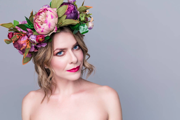 όμορφη νεαρή γυναίκα μοντέλο με φωτεινά λουλούδια στο κεφάλι της σε γκρι φόντο - Φωτογραφία, εικόνα