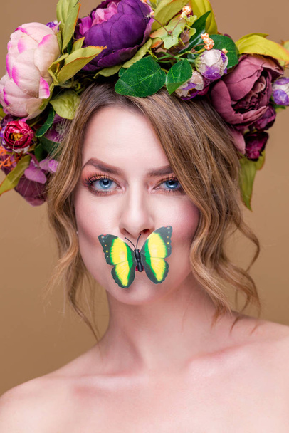 tender young woman posing in flower wreath with artificial  greenbutterfly,  isolated on ochet background. butterfly covering mouth of a young womans face - Zdjęcie, obraz
