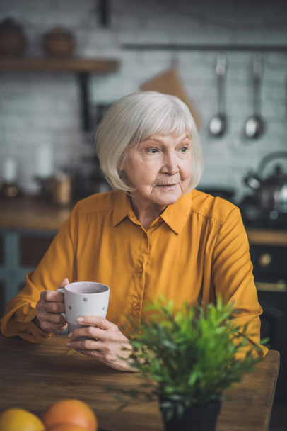 Good-looking elderly lady in yellow sitting in her kitchen - Фото, изображение