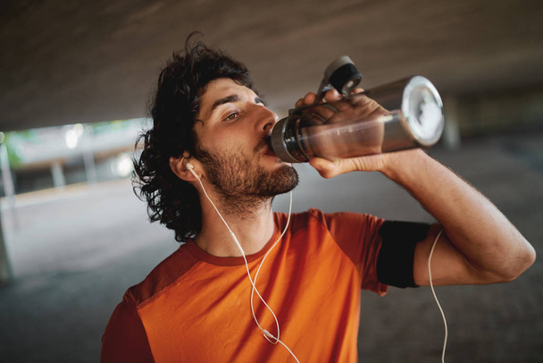 Close-up portrait of a handsome fit young man with earphones in his ears drinking water from a reusable water bottle - Foto, Bild