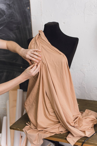 Το Seamstress δοκιμάζει ύφασμα σε μαύρη κούκλα σε εργαστήριο ραπτικής. Ο σχεδιαστής σχεδιάζει ένα φόρεμα από μπεζ ύφασμα και δημιουργεί ρούχα. θηλυκό μοδίστρα αποδίδουν ύφασμα σε κούκλα με βελόνες. κρε - Φωτογραφία, εικόνα