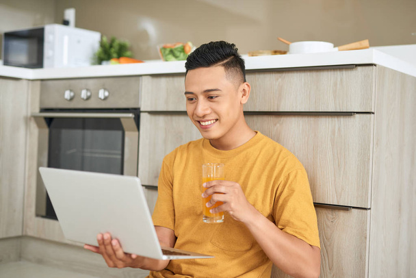 Jeune homme d'affaires sourit, prend son petit déjeuner et travaille sur un ordinateur portable dans une cuisine lumineuse
. - Photo, image