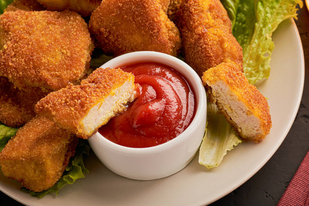 Nahaufnahme einer Scheibe Chicken Nuggets in Ketchup in einem Teller mit einem Bündel Nuggets auf dunklem Hintergrund - Foto, Bild