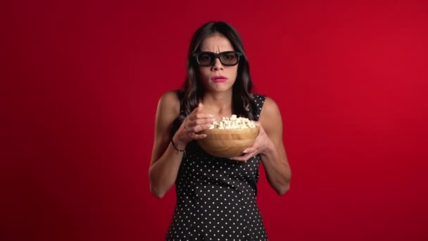 Young hispanic woman in 3d glasses watching horror movie and eating popcorn on red studio background. Frightened girl because of unexpectedness plot in film. - Záběry, video