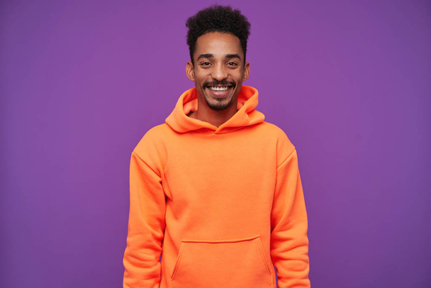 Cheerful young dark haired bearded brunette male with dark skin smiling pleasantly while standing over purple background with hands down, being in good mood - Fotó, kép