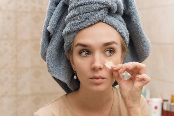 Ένα όμορφο κορίτσι με μια πετσέτα στο κεφάλι κολλάει υδρογέλη κάτω από τα μάτια της στο πρόσωπό της. Το κορίτσι φροντίζει το δέρμα και ενυδατώνει την περιοχή γύρω από τα μάτια. Θεραπείες κατ 'οίκον φροντίδας στο μπάνιο - Φωτογραφία, εικόνα