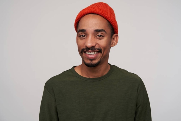 Portrait of beautiful young brown-eyed dark skinned male with beard looking cheerfully at camera with pleasant smile, dressed in red hat and khaki pullover over white background - Foto, Bild
