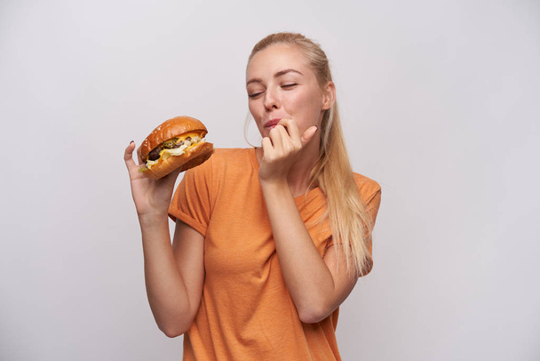 Красивая молодая симпатичная блондинка с поникшей прической, держащая в поднятой руке вкусный чизбургер и смакующая пальцы, стоящая на белом фоне - Фото, изображение