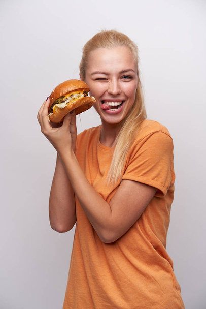 Funny shot of lovely young blonde woman with ponytail hairstyle giving wink to camera and showing her tongue while posing over white background with fresh burger in her hands - Photo, image