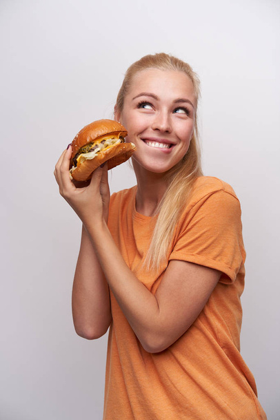 Vrolijke jonge vrij lange haren blonde dame met casual kapsel heffende handen met smakelijke hamburger en kijken gelukkig opzij met charmante glimlach, poseren over witte achtergrond - Foto, afbeelding