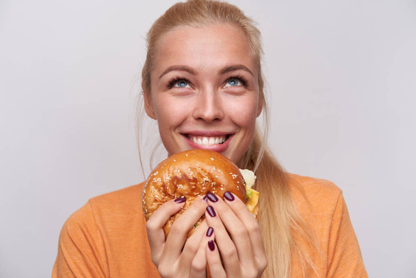 Закрытие жизнерадостной голубоглазой молодой блондинки с большим вкусным гамбургером, счастливо смотрящим вверх и широко улыбающимся, одетым в повседневную одежду, позируя на белом фоне - Фото, изображение