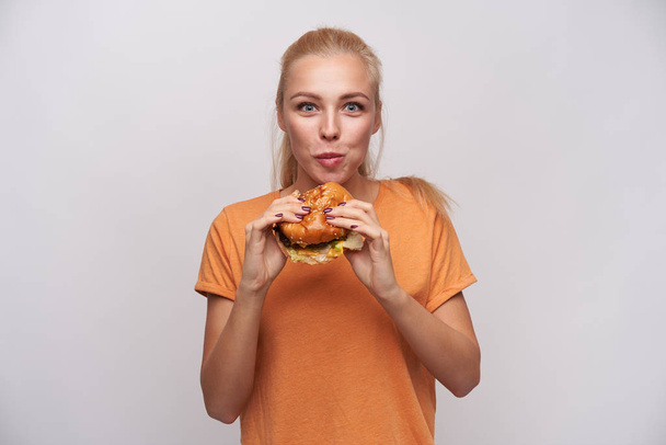 Indoor-Foto der attraktiven jungen hungrigen blonden Dame mit Pferdeschwanzfrisur kaut köstlichen Hamburger und schaut aufgeregt in die Kamera, gekleidet in orangefarbenem T-Shirt vor weißem Hintergrund - Foto, Bild