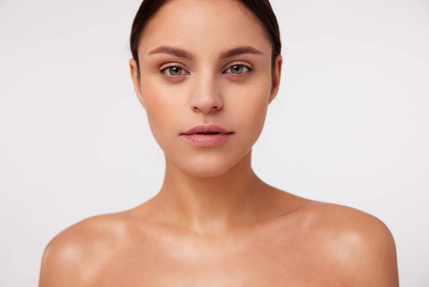 Close-up of beautiful young brunette green-eyed woman with natural makeup looking sincerely at camera, standing over white background with bare shoulders - Photo, Image