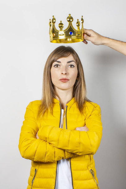 Giovane donna in un piumino giallo e una mano tiene una corona sopra la testa su uno sfondo chiaro. Concetto regina, ambizione, sogno, aspirazione, ego
 - Foto, immagini