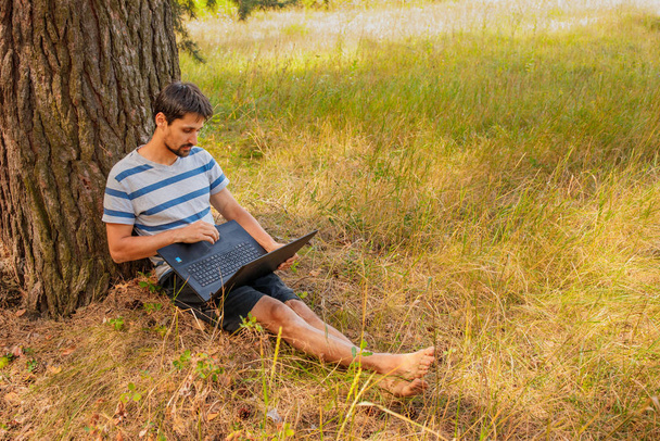 Ένας νεαρός μελαχρινός με γενειάδα κάθεται στο γρασίδι με ένα λάπτοπ. Φοιτητής εργάζεται σε εξωτερικούς χώρους - Φωτογραφία, εικόνα