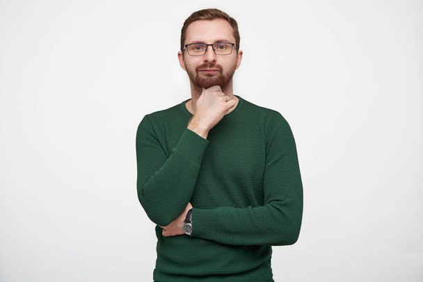 Portrait of thoughtful young brunette bearded man wearing glasses and green sweater while posing over white background, holding his chin with raised hand and keeping lips folded - Photo, Image