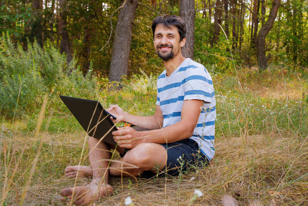 Молодой брюнетка с бородой сидит на траве с ноутбуком. Студенты работают на открытом воздухе
 - Фото, изображение