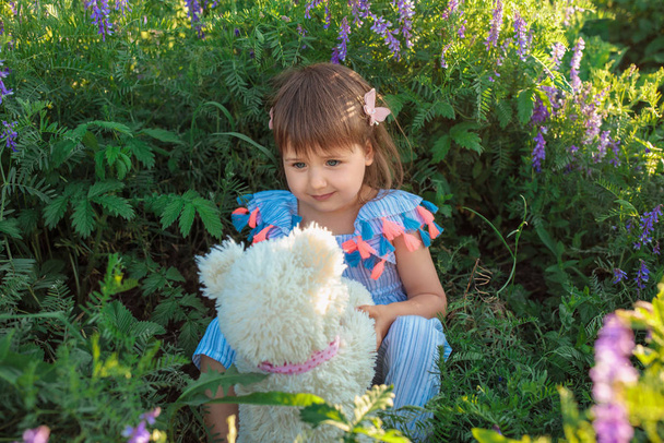 薄手のドレス姿で草の上に腰掛けている白人の少女は、テディベアを見ている。愛とケアを感じてください。家族の友情の概念. - 写真・画像