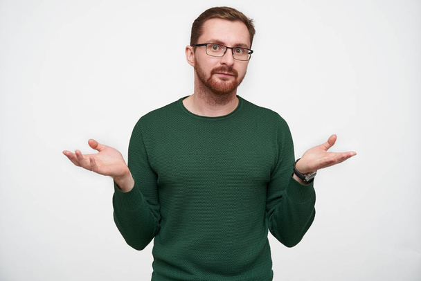 Puzzled young handsome brown haired bearded male shugging with raised palms and looking at camera with folded lips, wearing green pullover and eyewear over white background - Photo, Image