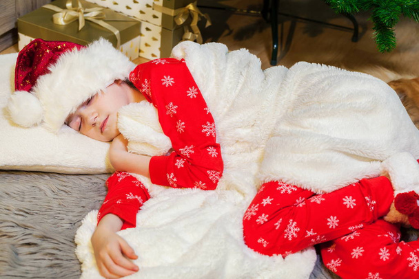 Το κοριτσάκι στην πιτζάμα αποκοιμήθηκε κοντά στο δέντρο την παραμονή των Χριστουγέννων. - Φωτογραφία, εικόνα