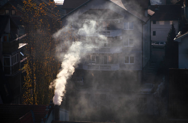 Smoking chimneys at roofs of houses emits smoke, smog at sunrise, pollutants enter atmosphere. Environmental disaster. Harmful emissions and exhaust gases into air. Fog, winter day, heating season. - Photo, Image