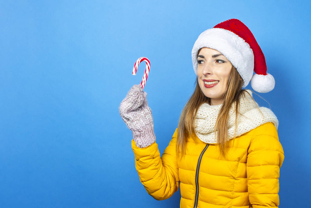 Young woman with a smile in a yellow jacket and a hat of Santa Claus holds a candy cane on a blue background. Concept of the winter holidays, Christmas, New Year, surprise, shock. Banner - Zdjęcie, obraz