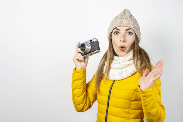 Young woman with a surprised face in a yellow down jacket and hat holds a camera in her hands on a light background. Concept of beauty and fashion, winter, autumn, technology. Banner - Photo, image