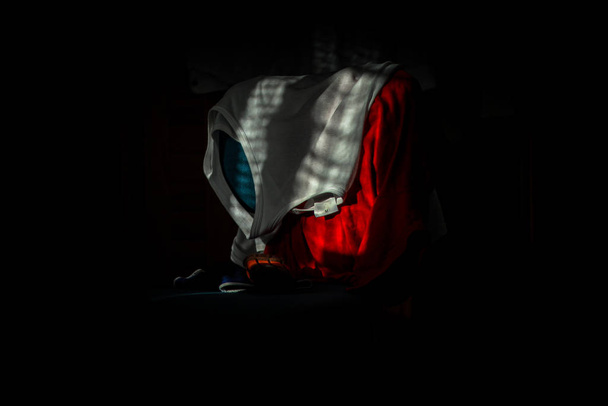 White undershirt thrown over red shirt and blue chair in dark room, lit by sun through blinds - 写真・画像