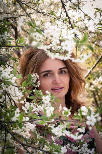 selective focus of beautiful woman with long hair standing behind tree with flowering cherry branches and looking at camera in garden - Foto, Imagem