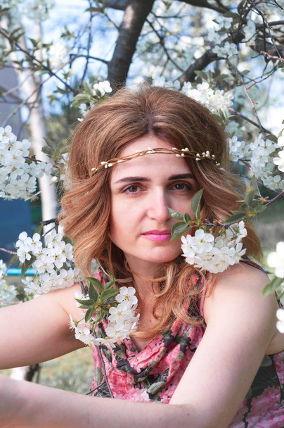 soft focus of attractive woman with long hair in floral wreath looking at camera surrounded by blooming cherry branches in garden - Foto, Imagem