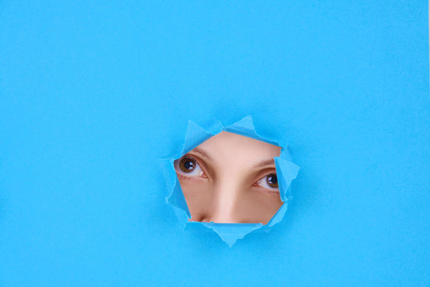 Μέρος του προσώπου μιας γυναίκας τα μάτια και η μύτη μιας ελκυστικής γυναίκας κοιτάζοντας στην κάμερα, πίσω από ένα σπασμένο τοίχο ή σχισμένο χαρτί, σε μπλε φόντο. Χώρος για κείμενο, επιστολόχαρτο ή φεμινιστικό σχεδιασμό - Φωτογραφία, εικόνα